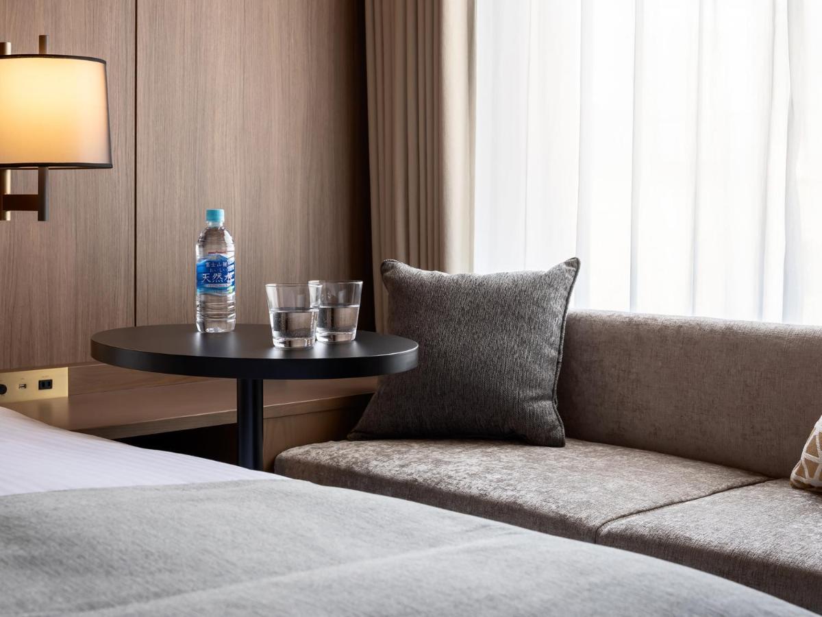 Mitsui Garden Hotel Roppongi Tokyo Premier Екстер'єр фото The photo depicts a cozy hotel room setting. On the left, there is a bedside table with a lamp. Against the wall, there is a comfortable-looking couch with a decorative pillow. In front of the couch, there's a small round table that holds a bottle of
