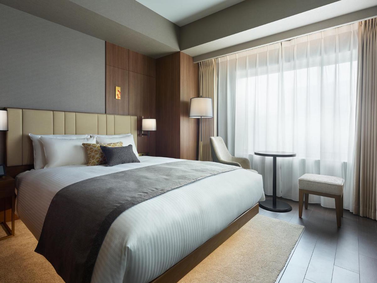 Mitsui Garden Hotel Roppongi Tokyo Premier Екстер'єр фото The photo shows a modern hotel room featuring a comfortable bed with white linens and a decorative throw pillow. The room has a neutral color palette with wooden accents on the walls. There are two bedside lamps and a chair positioned next to a small