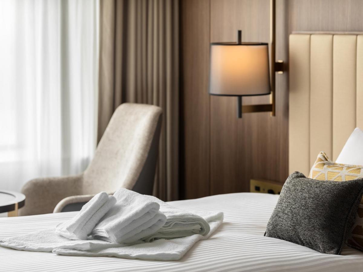 Mitsui Garden Hotel Roppongi Tokyo Premier Екстер'єр фото The image shows a stylish hotel room. In the foreground, there is a neatly made bed with white linens, featuring a stack of white towels arranged in a decorative manner. To the side, there is a gray upholstered armchair and a small table. The backgro