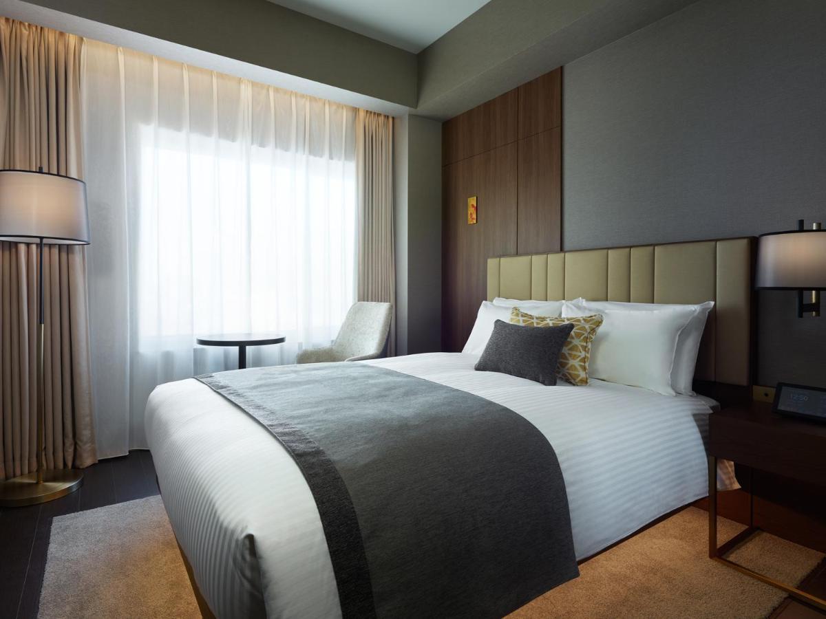 Mitsui Garden Hotel Roppongi Tokyo Premier Екстер'єр фото The photo shows a modern hotel room featuring a neatly made bed with white linens and a decorative gray throw blanket. To the side, there is a small round table and a comfortable chair, creating a cozy seating area. The room is well-lit with natural 