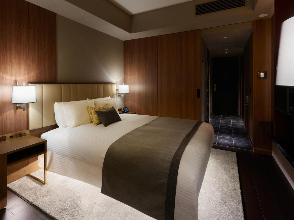Mitsui Garden Hotel Roppongi Tokyo Premier Екстер'єр фото The image shows a modern hotel room with a warm and elegant design. In the center, there is a neatly made bed with white linens and a dark gray throw blanket. Flanking the bed are two bedside lamps providing soft lighting. The walls have a rich wood 