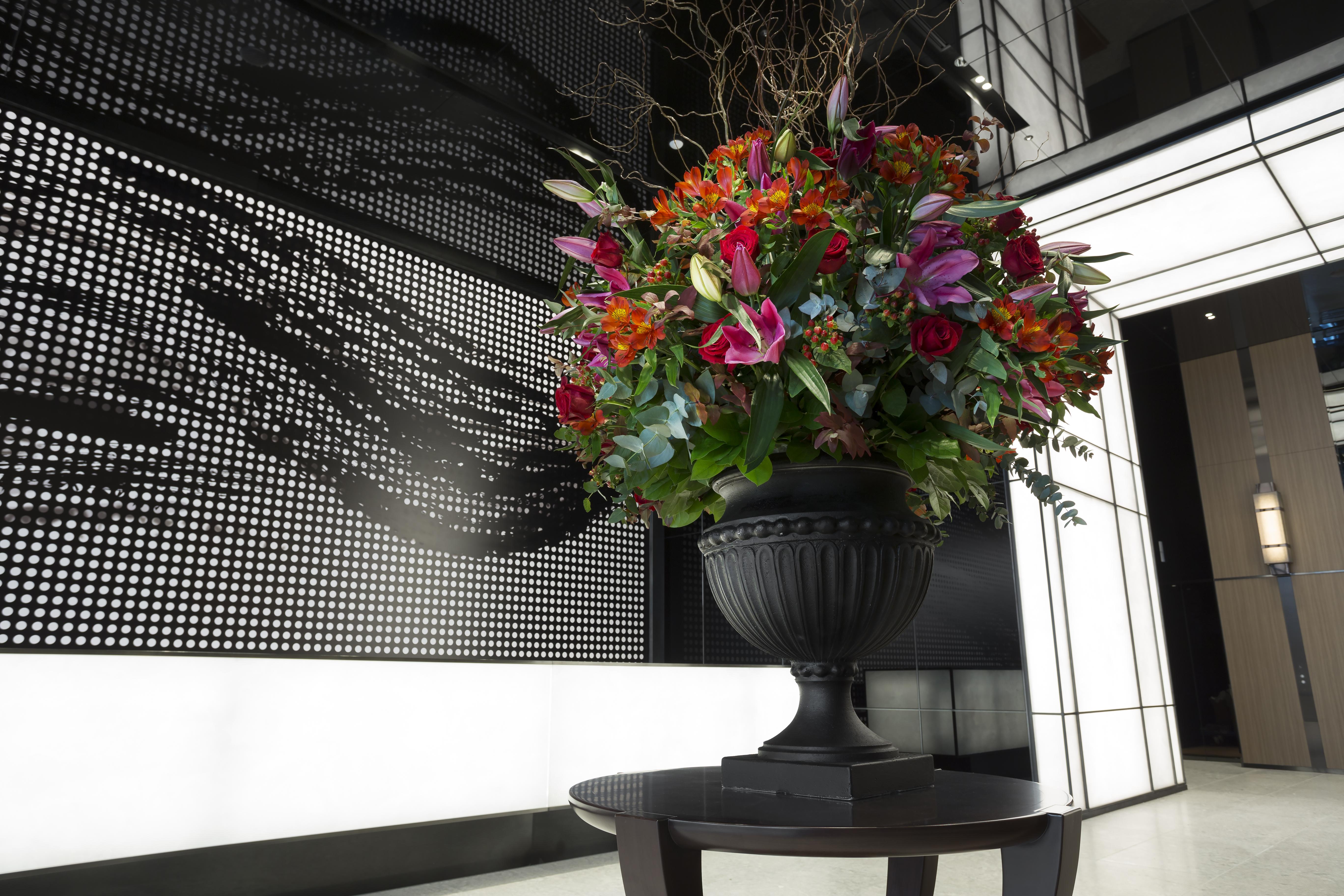 Mitsui Garden Hotel Roppongi Tokyo Premier Екстер'єр фото The photo features a large, vibrant floral arrangement in an ornate black urn. The arrangement is composed of various colorful flowers and greenery, showcasing shades of red, pink, orange, and green. This floral display is positioned on a small table