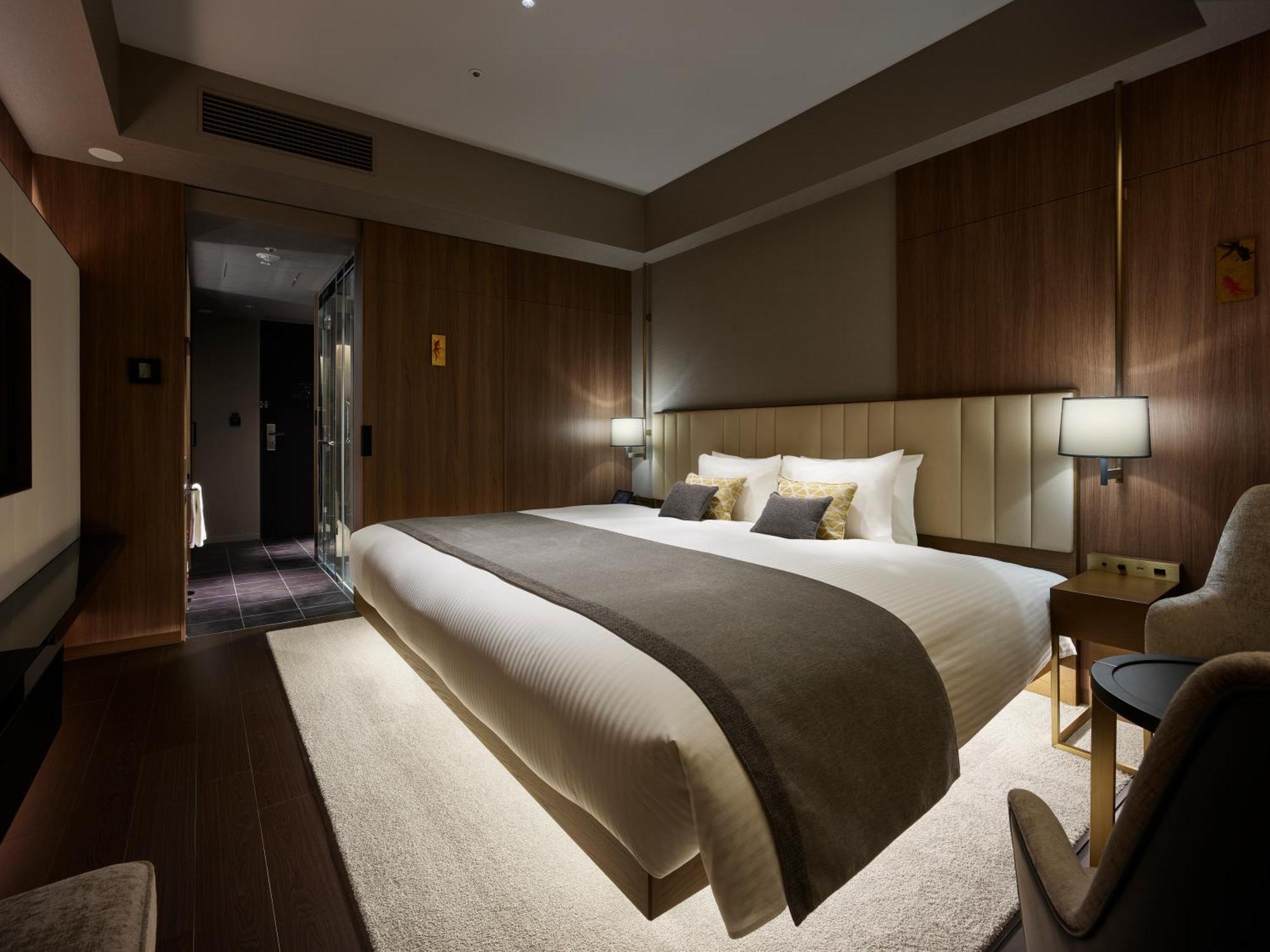 Mitsui Garden Hotel Roppongi Tokyo Premier Екстер'єр фото The photo shows a modern hotel room featuring a large bed with a neutral-colored duvet and a dark throw blanket. The headboard is upholstered, and there are two bedside lamps on either side of the bed. The walls have a warm wood paneling, contributin