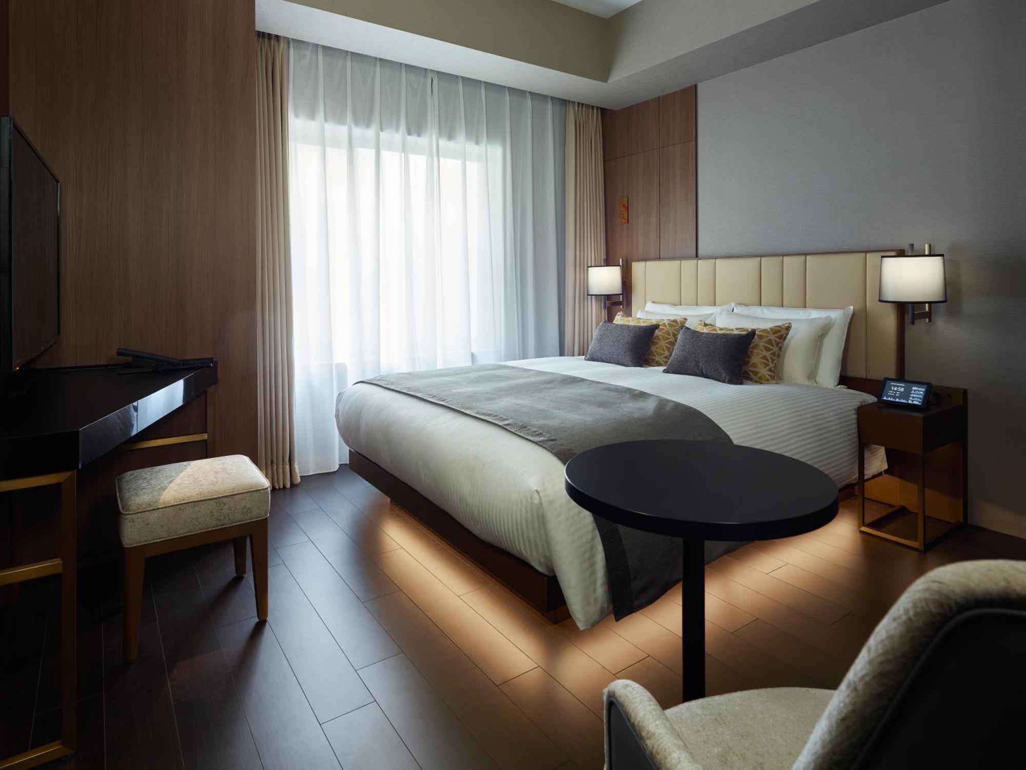 Mitsui Garden Hotel Roppongi Tokyo Premier Екстер'єр фото The photo shows a modern hotel room featuring a large bed with decorative pillows. There are large windows with sheer curtains allowing natural light to filter in. To one side, there is a sleek desk with a chair and a small round table in the center 
